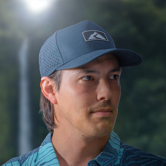 man wearing a blue performance hat with a rubber patch with the Waiākea logo on it with a floral background behind him