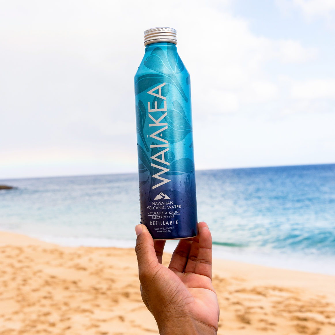Hand holding a Waiakea bottle on a beach with ocean and sky in the background