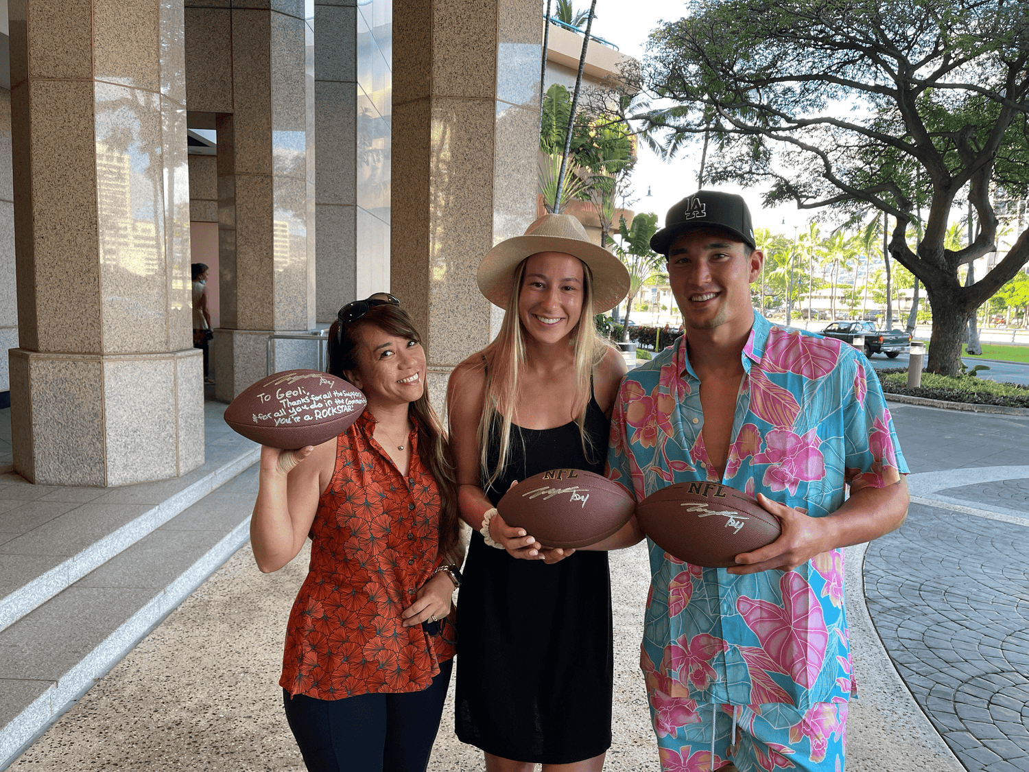 Three people holding signed footballs outdoors.