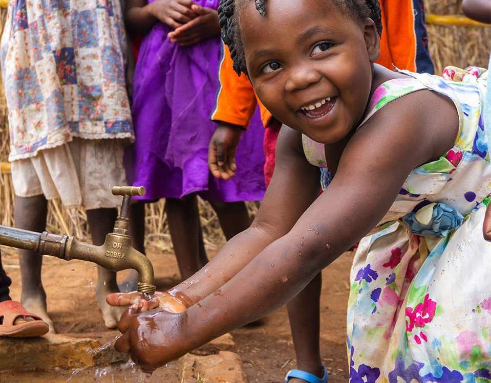 Child washing hands at outdoor water faucet, smiling.
