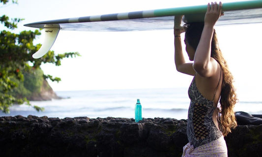 Woman carrying surfboard on head with a bottle of Waiakea in the background