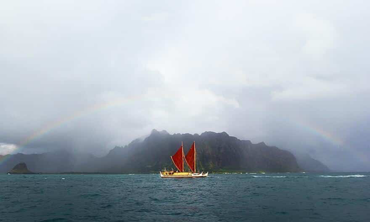 Hōkūle‘a passing in front of a mountain with a rainbow overhead