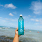 Person holding a Waiakea water bottle with a beach and ocean in the background