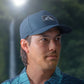 man wearing a blue performance hat with a rubber patch with the Waiākea logo on it with a floral background behind him