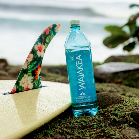 Waiakea water bottle on a surfboard with a floral fin, set against a beach backdrop.