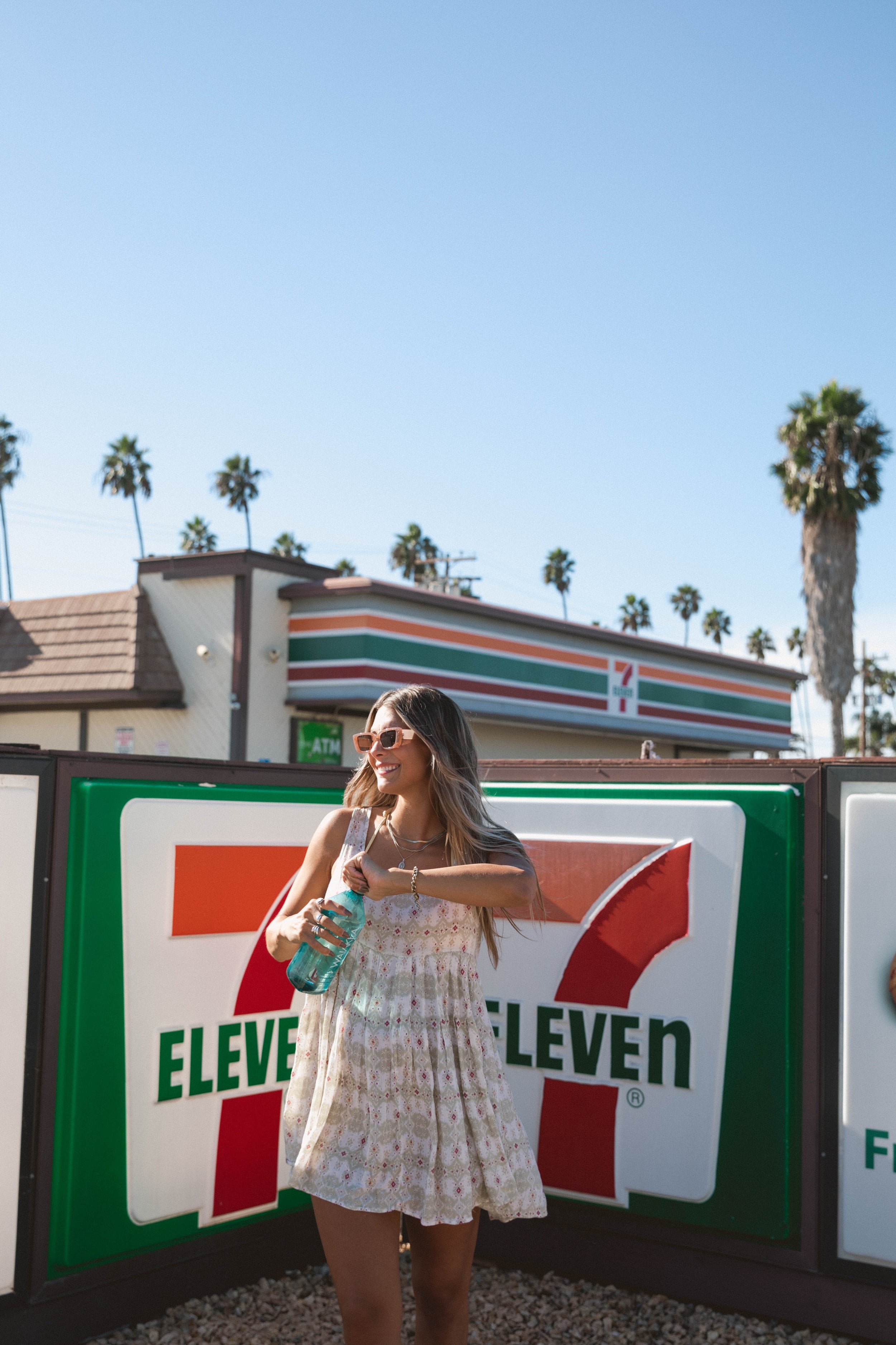 Woman holding a drink in front of a 7-Eleven sign with palm trees in the background.